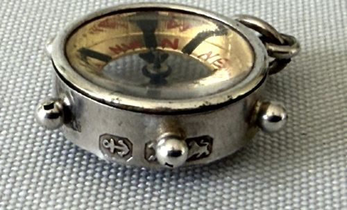 A miniature silver fob compass designed in the shape of a ships wheel with a central directional needle and suspension ring. View III