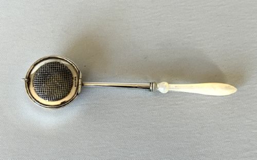 fig 1. A late Victorian silver tea infuser featuring a circular mesh cage connected to a stem with a light-coloured carved handle.