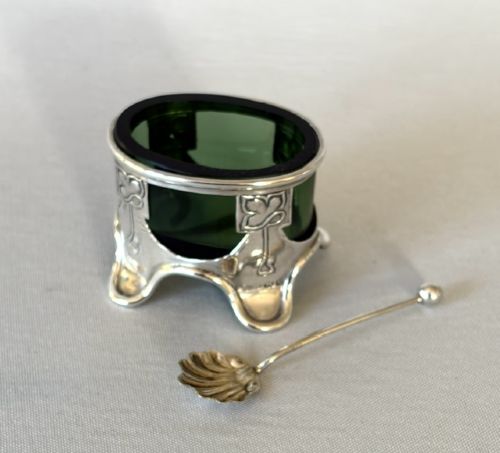 fig 1. An Art Nouveau silver salt cellar frame featuring stylized organic motifs and resting on curved feet, housing a fitted green glass liner with an accompanying silver spoon.