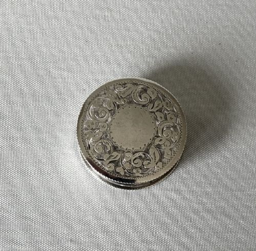 fig 1. A small circular silver box featuring hand-engraved foliage and scroll patterns surrounding an unengraved circular centre.