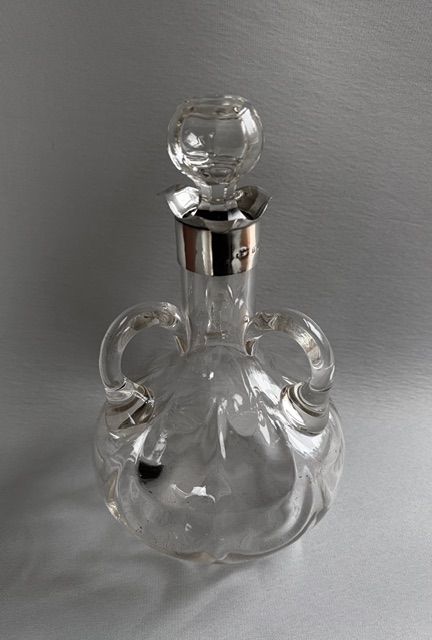 fig 1. An Edwardian clear glass decanter featuring a bulbous body, three loop handles, a hallmarked sterling silver collar, and a circular glass stopper.