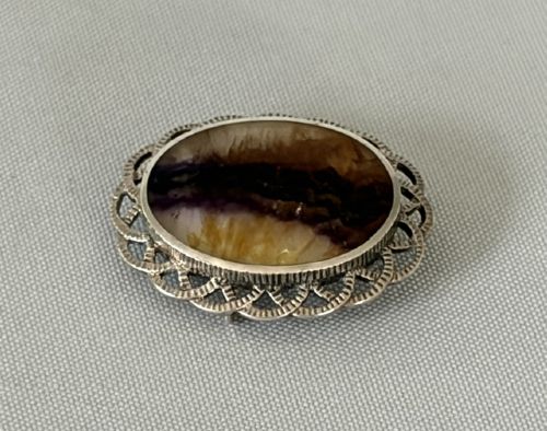 fig 1. An oval Art Deco silver brooch featuring a polished Blue John stone with purple and yellow banding, encircled by a textured scalloped lattice border.
