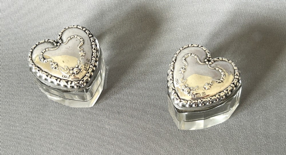 A pair of heart-shaped clear cut glass vanity jars featuring chased sterling silver lids with floral motifs.