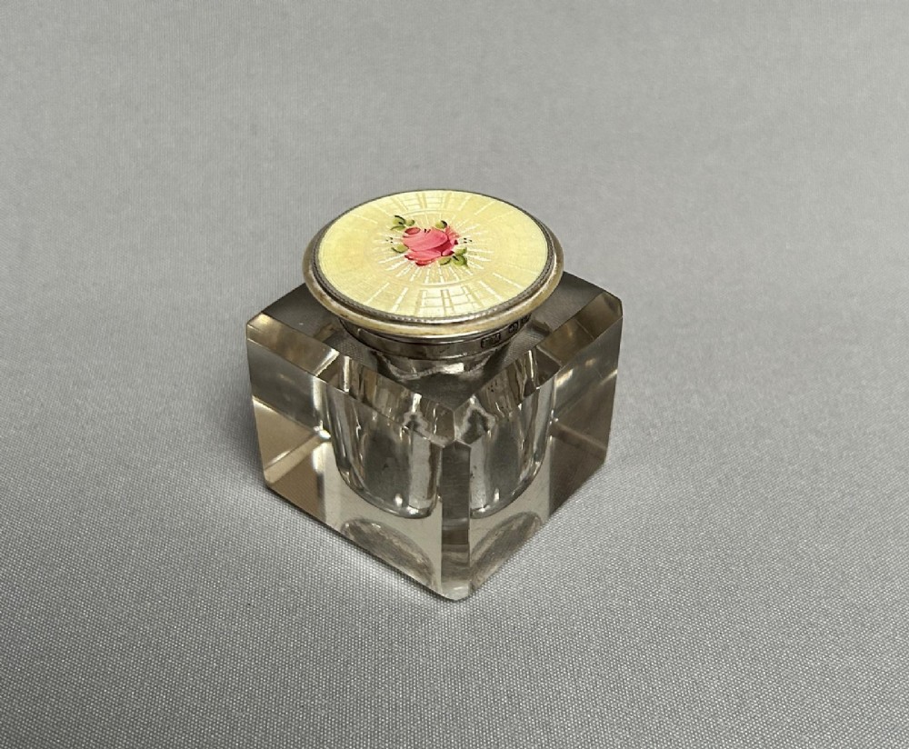 A heavy square glass inkwell with a hallmarked silver collar and a pale yellow guilloche enamel lid featuring a hand-painted pink rose motif.
