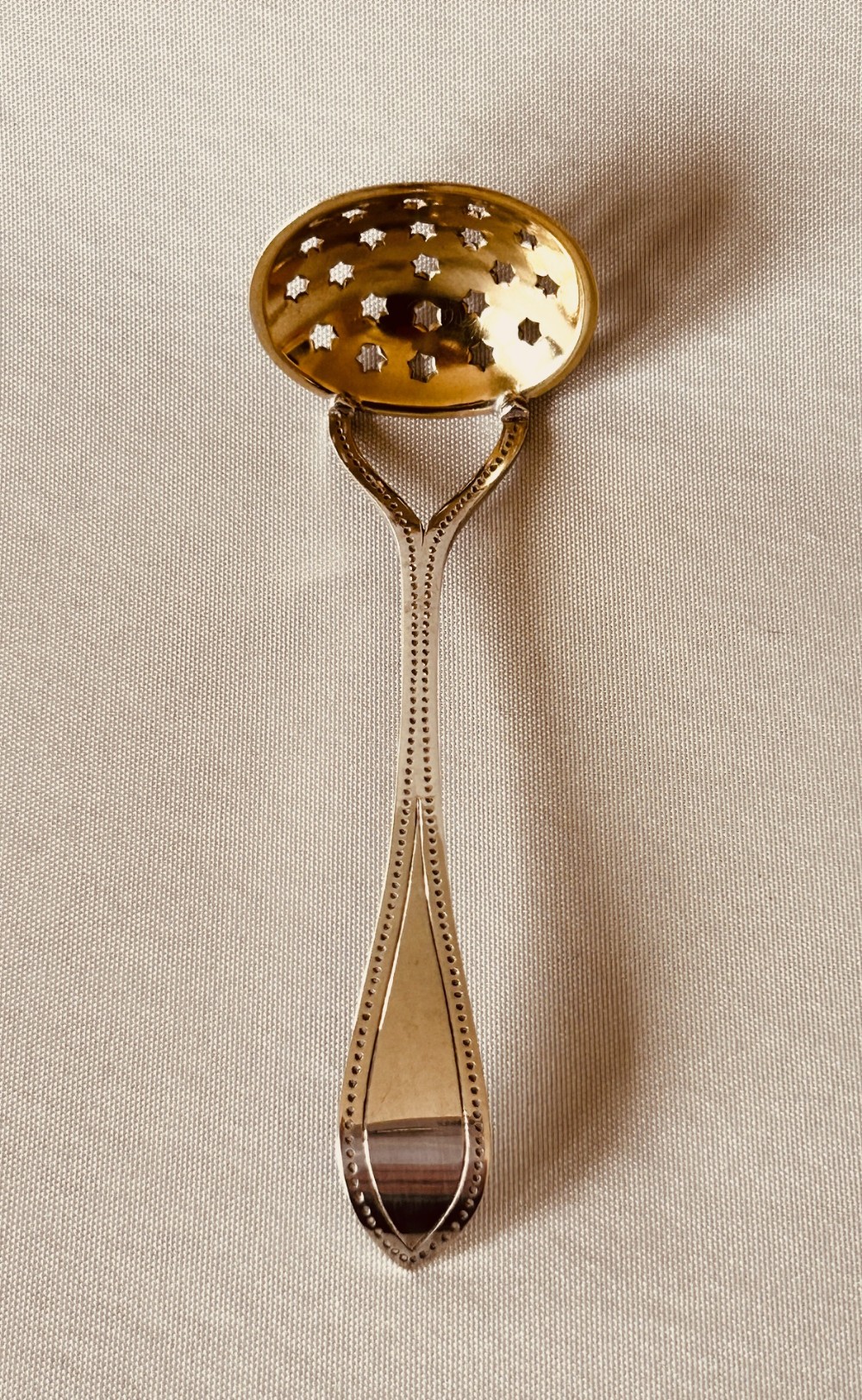 An Arts and Crafts silver gilt sifter spoon featuring star-shaped perforations in an oval bowl and a beaded edge handle.