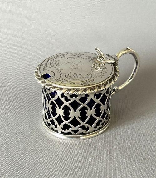 An early Victorian silver mustard pot with pierced fretwork sides, a cobalt blue glass liner, an engraved flat hinged lid, and a scroll handle.