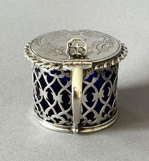 An early Victorian silver mustard pot with pierced fretwork sides, a cobalt blue glass liner, an engraved flat hinged lid, and a scroll handle. View IV