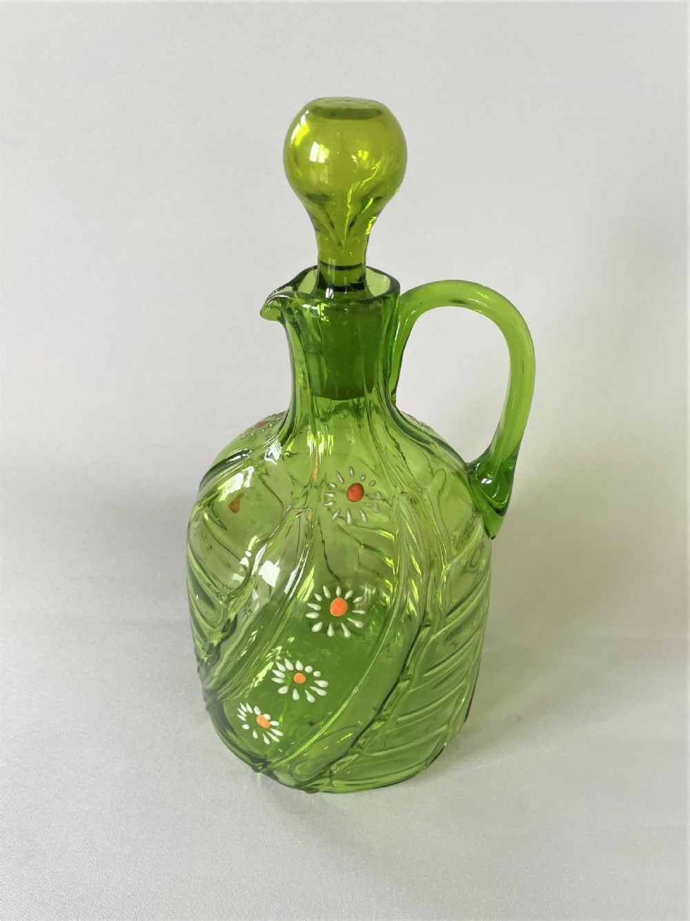 A Victorian green glass wine jug featuring hand-painted white enamel daisy motifs, a swirled optic body, a loop handle, and a matching solid glass stopper.
