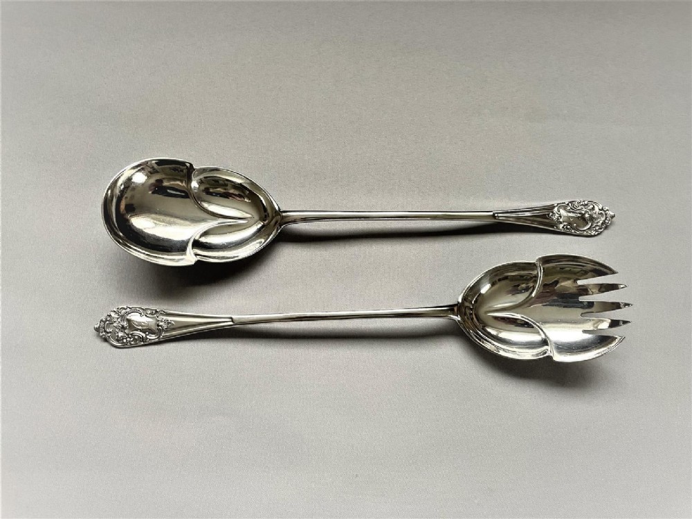 A pair of Edwardian silver salad servers consisting of a spoon and a fork with stylized leaf-shaped bowls and scroll-embossed handle terminals.