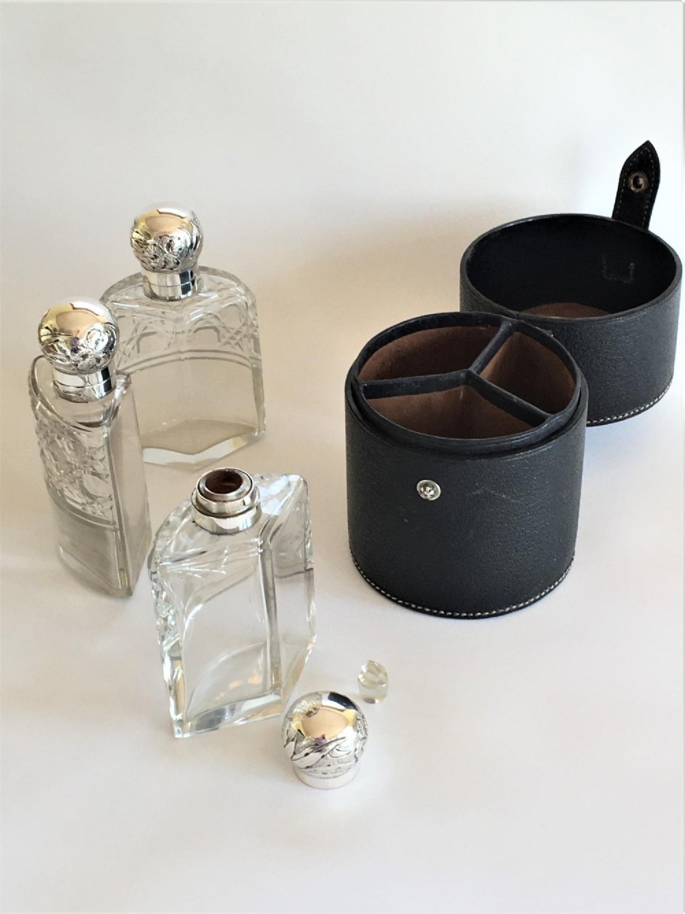 Three wedge-shaped clear glass cologne bottles with silver caps resting beside an open cylindrical black leather travel case.