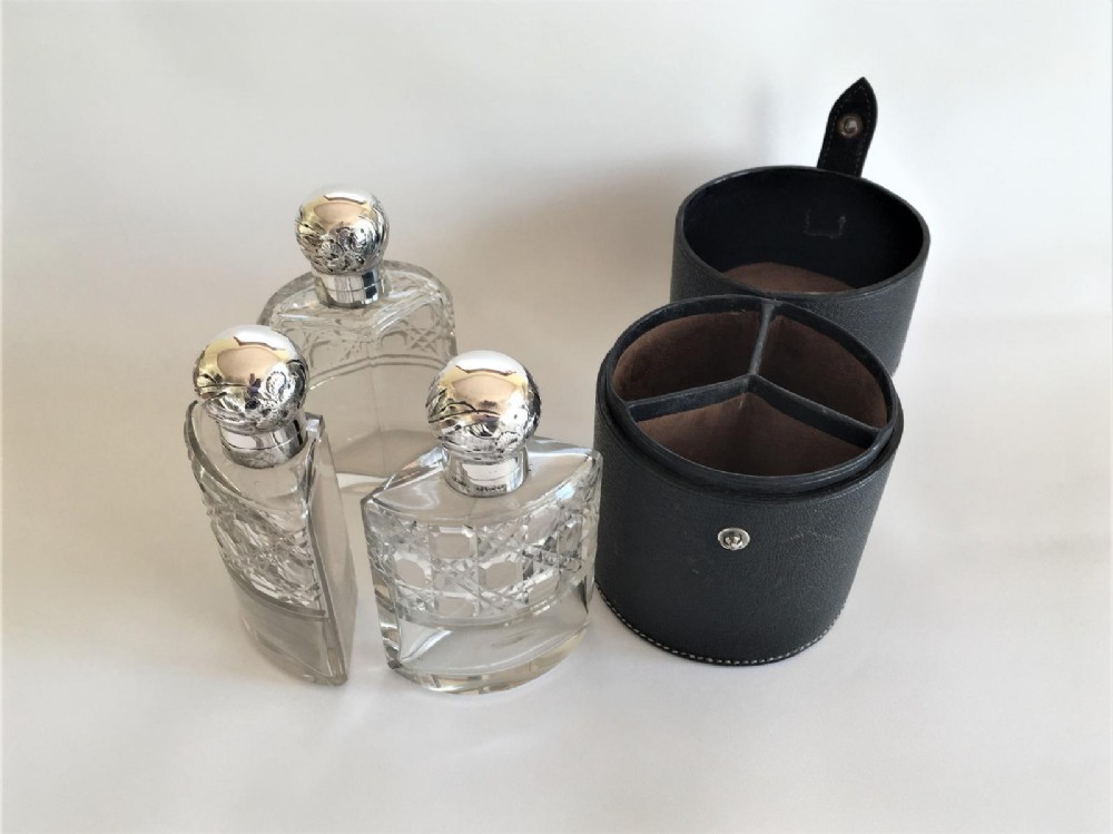 Three wedge-shaped clear glass cologne bottles with silver caps resting beside an open cylindrical black leather travel case. View III