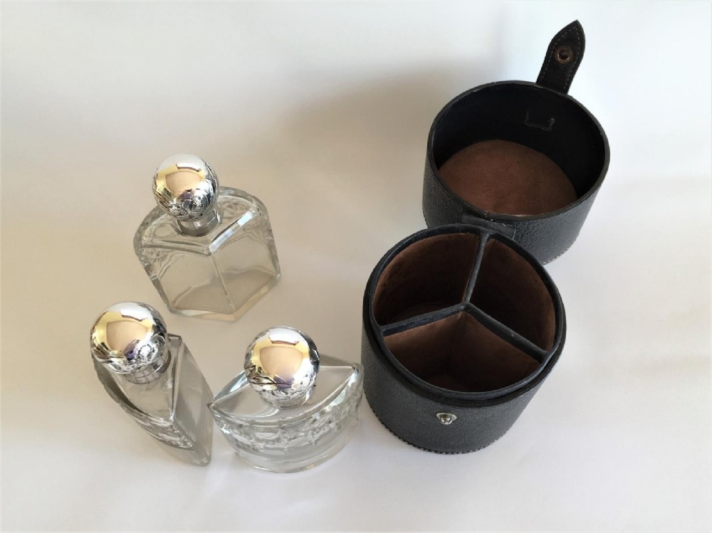 Three wedge-shaped clear glass cologne bottles with silver caps resting beside an open cylindrical black leather travel case. View VII