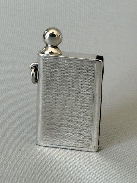 An Art Deco silver pocket lighter shaped like a small book with engine-turned textured engraving, a side attachment ring, and a spherical top screw cap.
