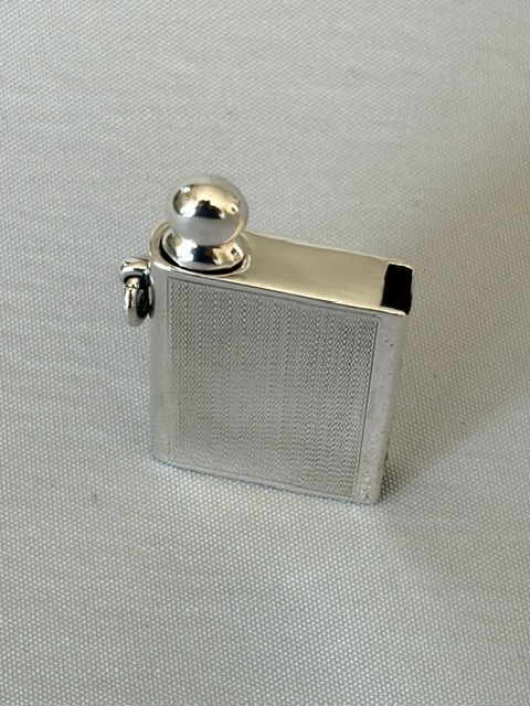 An Art Deco silver pocket lighter shaped like a small book with engine-turned textured engraving, a side attachment ring, and a spherical top screw cap. View II
