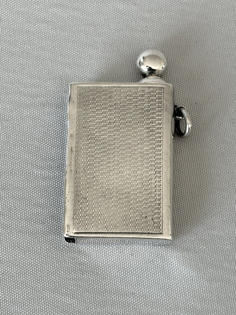 An Art Deco silver pocket lighter shaped like a small book with engine-turned textured engraving, a side attachment ring, and a spherical top screw cap. View III