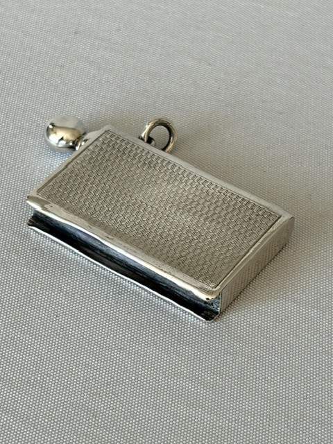 An Art Deco silver pocket lighter shaped like a small book with engine-turned textured engraving, a side attachment ring, and a spherical top screw cap. View IV
