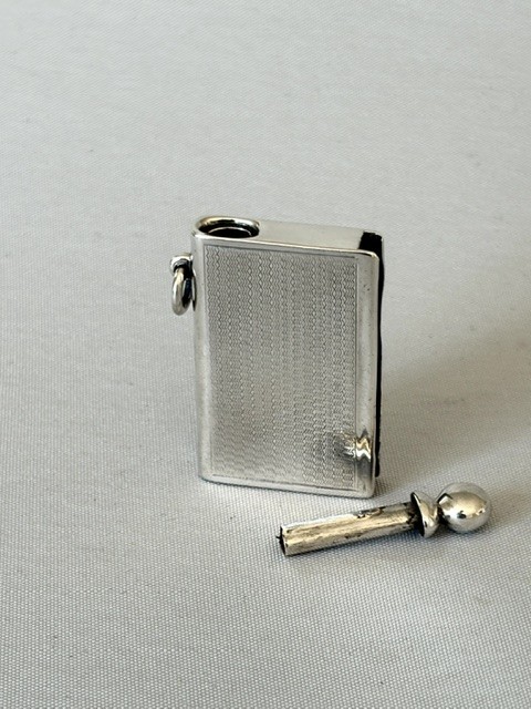 An Art Deco silver pocket lighter shaped like a small book with engine-turned textured engraving, a side attachment ring, and a spherical top screw cap. View V