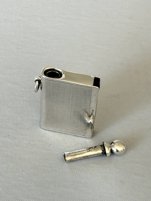 An Art Deco silver pocket lighter shaped like a small book with engine-turned textured engraving, a side attachment ring, and a spherical top screw cap. View VI