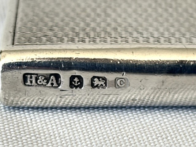 An Art Deco silver pocket lighter shaped like a small book with engine-turned textured engraving, a side attachment ring, and a spherical top screw cap. View VII
