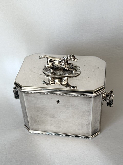 A silver-plated rectangular box with canted corners, featuring a horse finial on the lid, horse head handles on the sides, and a central keyhole. View II