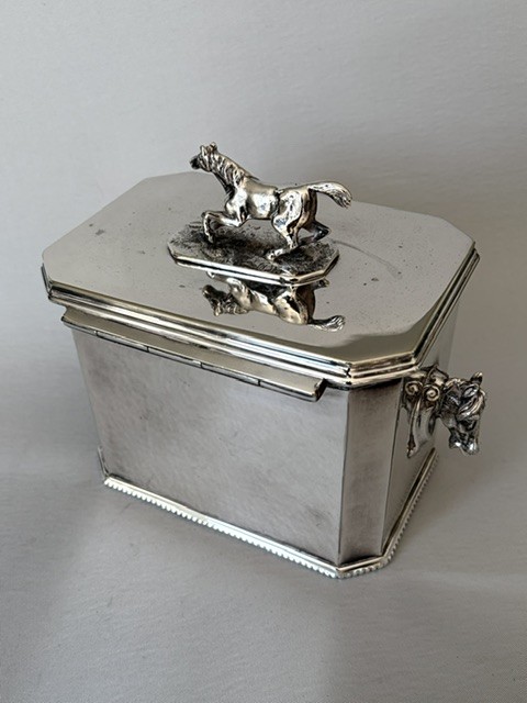 A silver-plated rectangular box with canted corners, featuring a horse finial on the lid, horse head handles on the sides, and a central keyhole. View VI