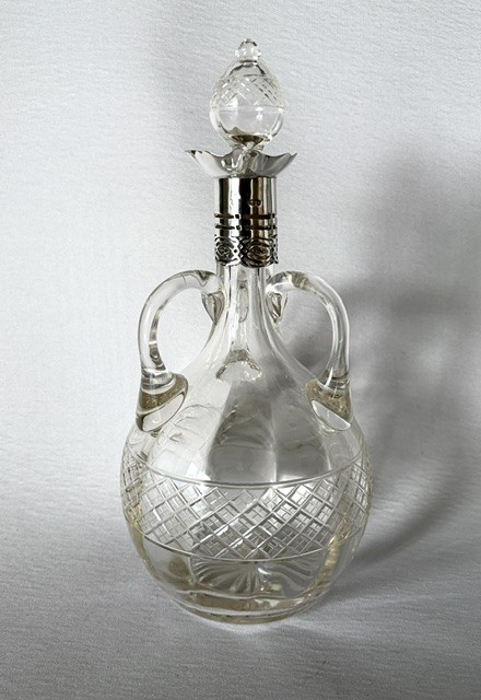 An Edwardian clear cut glass decanter featuring double scroll handles, a diamond-cut lower body, and a hallmarked silver collar topped with a faceted glass stopper.