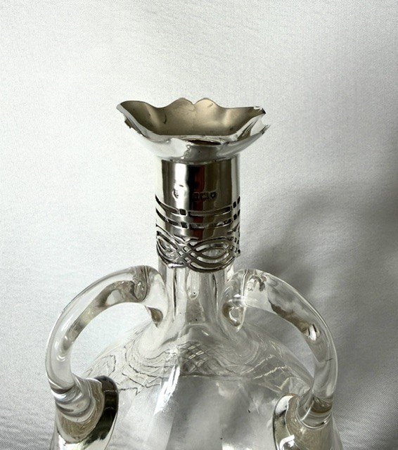 An Edwardian clear cut glass decanter featuring double scroll handles, a diamond-cut lower body, and a hallmarked silver collar topped with a faceted glass stopper. View IV