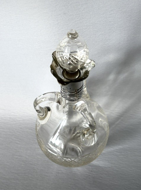An Edwardian clear cut glass decanter featuring double scroll handles, a diamond-cut lower body, and a hallmarked silver collar topped with a faceted glass stopper. View VI