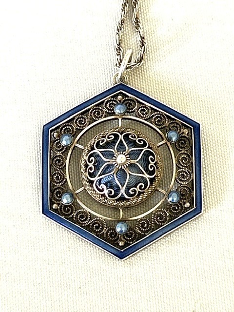 A hexagonal silver pendant with delicate wire filigree scrolls surrounding a central blue guilloche enamel dome, suspended from a silver chain.
