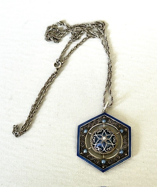 A hexagonal silver pendant with delicate wire filigree scrolls surrounding a central blue guilloche enamel dome, suspended from a silver chain. View II