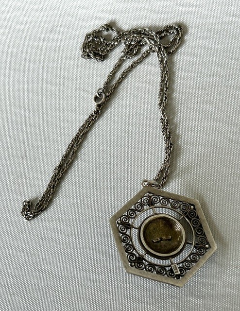 A hexagonal silver pendant with delicate wire filigree scrolls surrounding a central blue guilloche enamel dome, suspended from a silver chain. View III