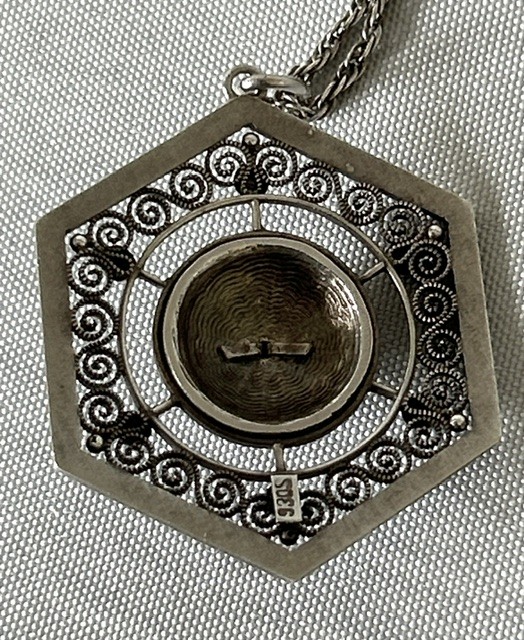 A hexagonal silver pendant with delicate wire filigree scrolls surrounding a central blue guilloche enamel dome, suspended from a silver chain. View IV