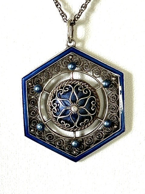 A hexagonal silver pendant with delicate wire filigree scrolls surrounding a central blue guilloche enamel dome, suspended from a silver chain. View V