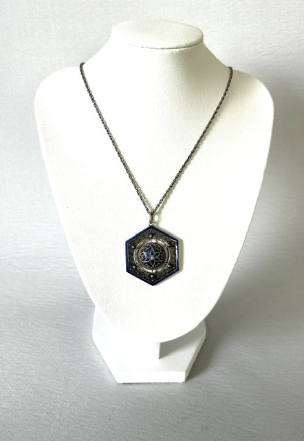 A hexagonal silver pendant with delicate wire filigree scrolls surrounding a central blue guilloche enamel dome, suspended from a silver chain. View VI