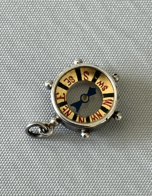A miniature silver fob compass designed in the shape of a ships wheel with a central directional needle and suspension ring. View VI