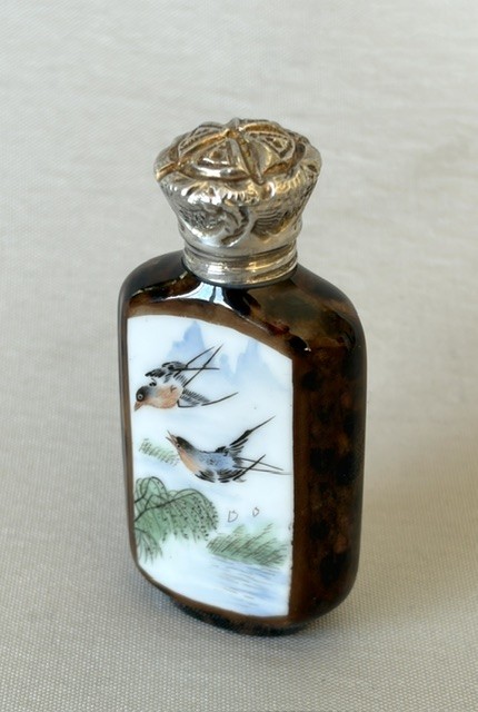 A small flattened oval scent bottle featuring a mottled brown body, a white enamel panel with a hand-painted bird among reeds, and a embossed silver cap. View II