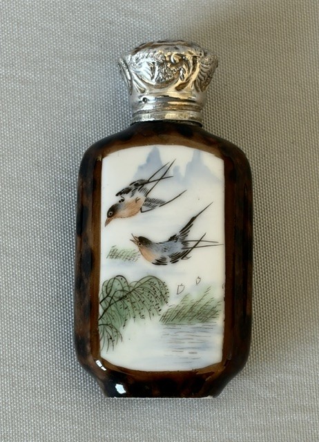 A small flattened oval scent bottle featuring a mottled brown body, a white enamel panel with a hand-painted bird among reeds, and a embossed silver cap. View IV