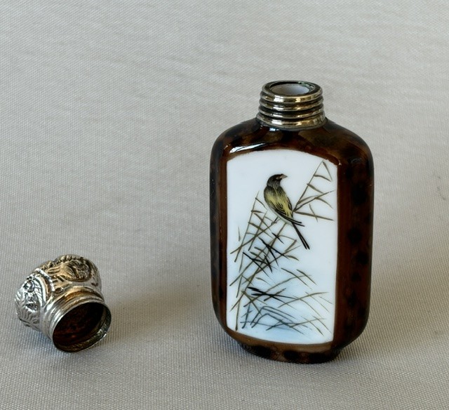 A small flattened oval scent bottle featuring a mottled brown body, a white enamel panel with a hand-painted bird among reeds, and a embossed silver cap. View V