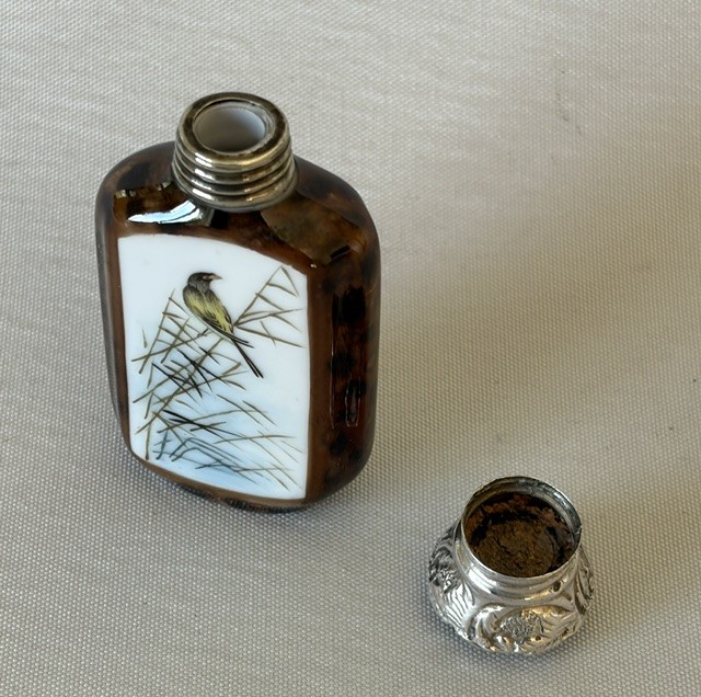 A small flattened oval scent bottle featuring a mottled brown body, a white enamel panel with a hand-painted bird among reeds, and a embossed silver cap. View VI