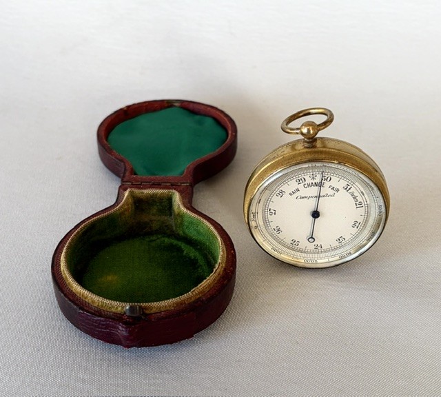 An Edwardian brass pocket barometer and altimeter displayed inside an open plush-lined maroon leather case. View III