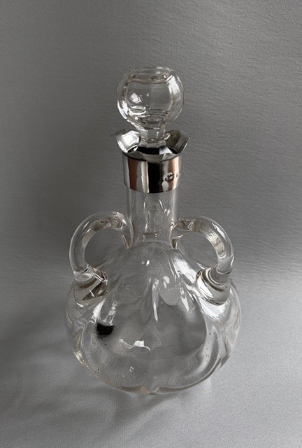 An Edwardian clear glass decanter featuring a bulbous body, three loop handles, a hallmarked sterling silver collar, and a circular glass stopper. View II