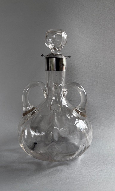An Edwardian clear glass decanter featuring a bulbous body, three loop handles, a hallmarked sterling silver collar, and a circular glass stopper. View III