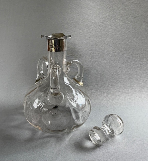 An Edwardian clear glass decanter featuring a bulbous body, three loop handles, a hallmarked sterling silver collar, and a circular glass stopper. View V
