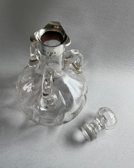 An Edwardian clear glass decanter featuring a bulbous body, three loop handles, a hallmarked sterling silver collar, and a circular glass stopper. View VI