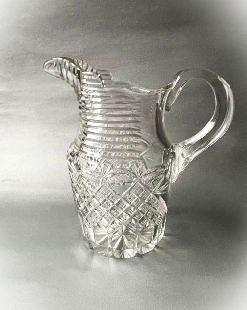A clear cut-glass water jug featuring a loop handle, horizontal step cuts around the neck, and diamond facets across the lower body.
