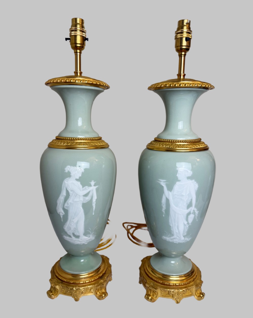 A pair of urn-shaped celadon green porcelain table lamps decorated with white classical figures in low relief, set upon ornate gilded bronze bases. View X