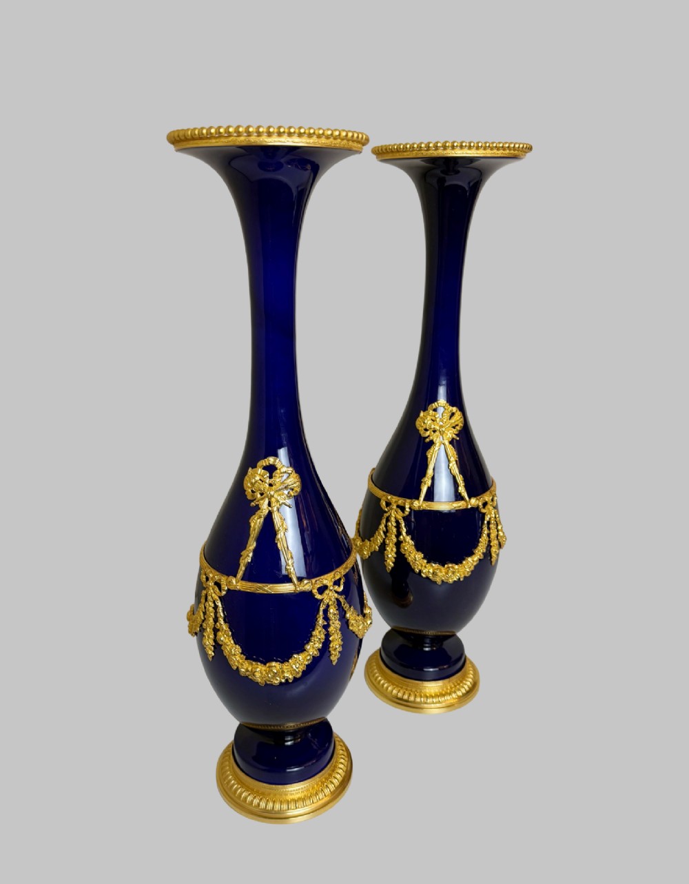 A pair of slender 19th-century cobalt blue porcelain vases featuring gilt bronze beaded rims, floral garland mounts, and circular cast pedestals. View II