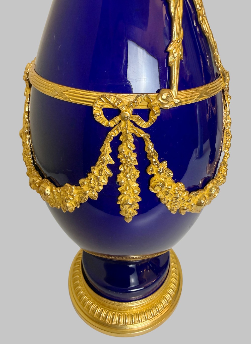 A pair of slender 19th-century cobalt blue porcelain vases featuring gilt bronze beaded rims, floral garland mounts, and circular cast pedestals. View VI