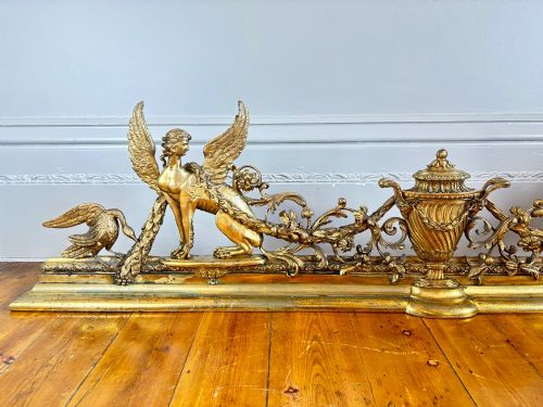fig 1. A long gilt bronze fireplace fender featuring winged sphinxes on either side, foliate scrollwork, and a central classical urn on a stepped base.