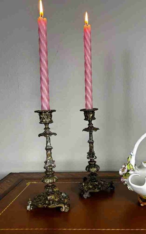 A pair of ornate 19th-century Regency brass candlesticks featuring detailed relief moulding and scrollwork bases holding lit candles.